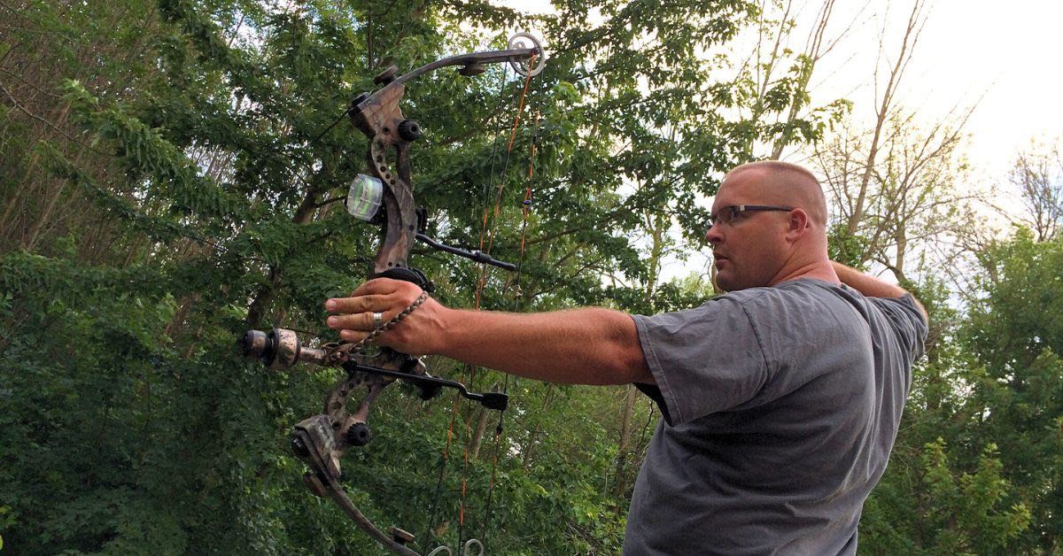 Bow hunter standing with compound bow drawn back with trees in the background.