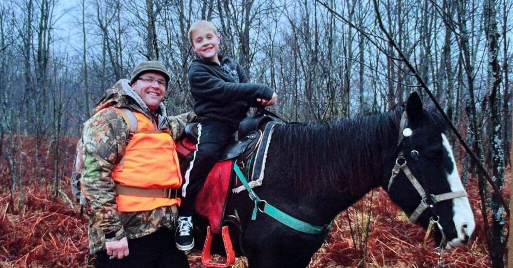 Hunter standing next to horse with son in the horse saddle