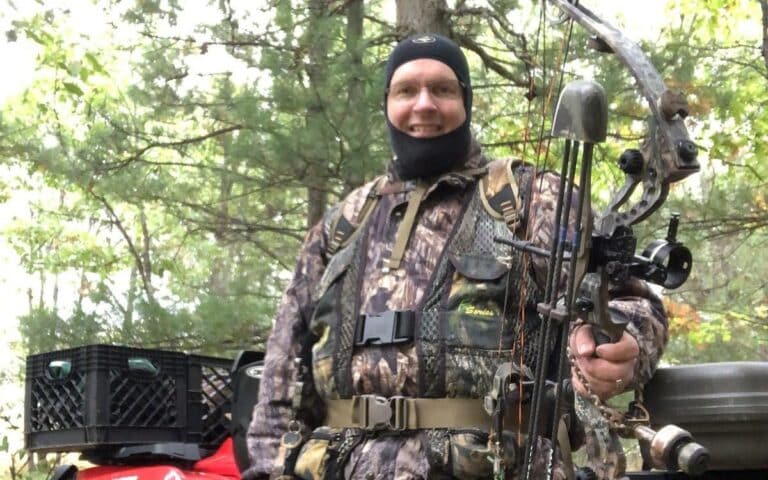 Bow hunter with safety harness and compound bow standing in front of a 4-wheelter