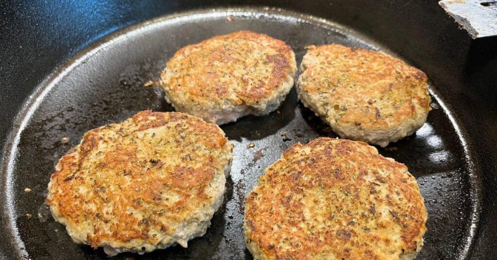 Four turkey burgers grilling in iron skillet on stovetop made with ground turkey and 5 ingredients