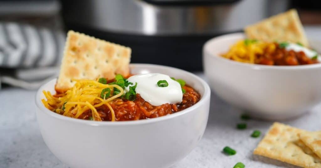 Two small white bowls with chili topped with shredded cheese, sour cream and crackers