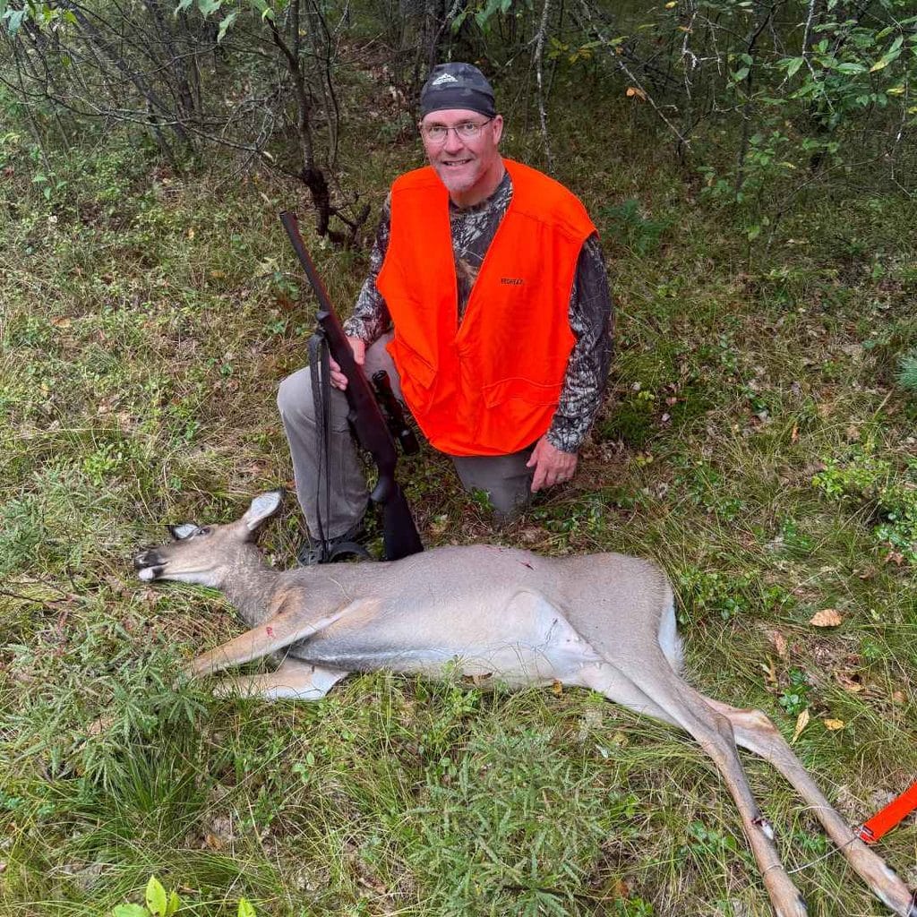 Hunter in blaze orange holding a rifle, kneeling beside a harvested doe in the Michigan woods.